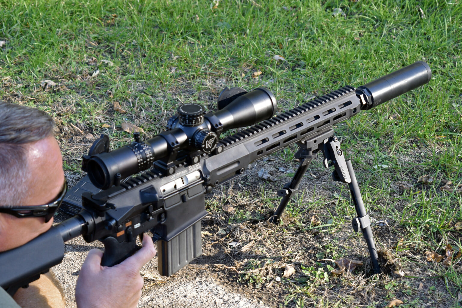 Over-the-should view of a white man in sunglasses lying on the ground, aiming a high-powered automatic rifle the suppressor attached.