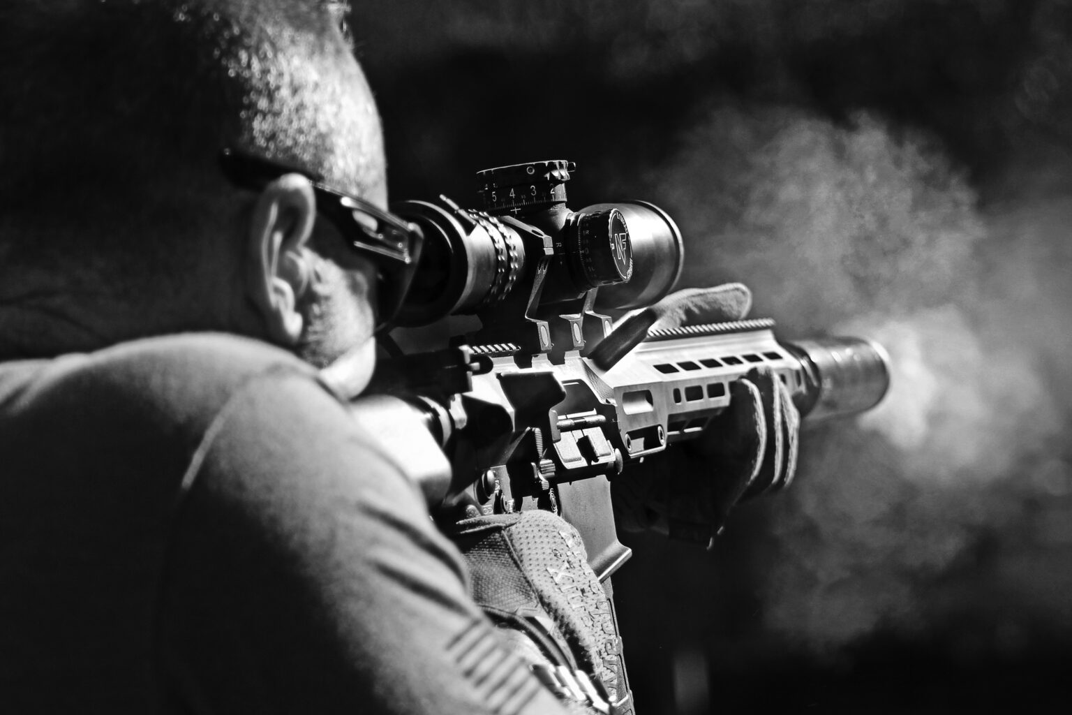 Black and white over-the-should view of man in sunglasses firing a high-powered automatic rifle.